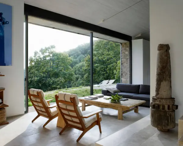 Salón moderno con sofás, sillones y mesa de centro rústica. Los suelos de piedra, con un estilo que evoca los años 70, contrastan con la vista exterior de un bosque frondoso.