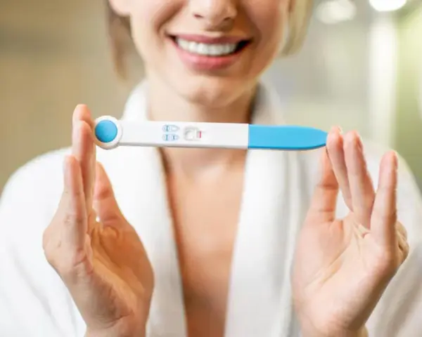 Uśmiechnięta kobieta w szlafroku trzyma test ciążowy z dwoma kreskami. To moment, kiedy warto zastanowić się, kiedy zrobić test ciążowy.