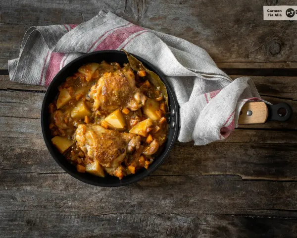 Estofado de pollo con patatas y zanahorias en sartén sobre mesa de madera.