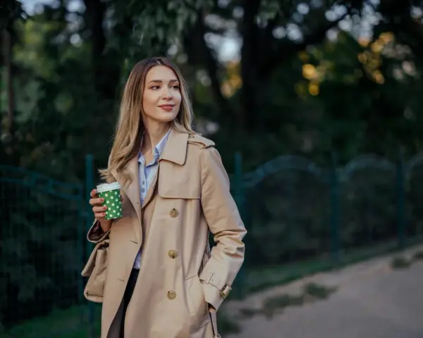 Donna con trench beige, un classico del marchio Burberry, sorseggia un caffè all'aperto.