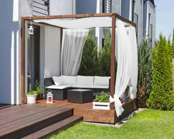 Drewniana pergola z białymi zasłonami to idealne miejsce na tarasie. Zasłony powieszone na drewnianej konstrukcji tworzą przytulną atmosferę.