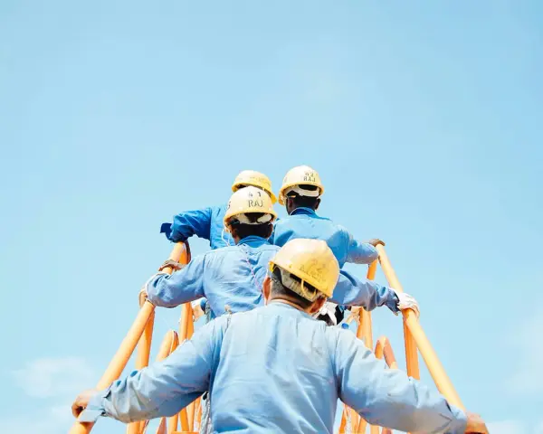 Trabajadores con cascos amarillos suben una escalera naranja contra un cielo azul claro. Podría ser una escena de cesión ilegal de trabajadores.