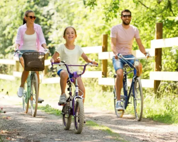 Rodzina cieszy się rodzinnym wypadem na **Mazury rowerem**. Radosna dziewczynka jedzie na fioletowym rowerze, a za nią rodzice.