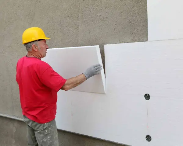 Operaio con casco giallo applica pannelli isolanti bianchi. Attenzione: il polistirolo è infiammabile.
