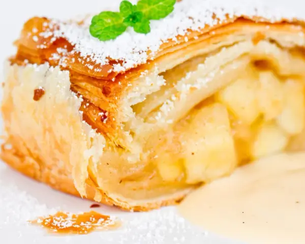 Ein Stück Apfelstrudel mit Filoteig, bestäubt mit Puderzucker und garniert mit Minze, serviert mit Vanillesauce.
