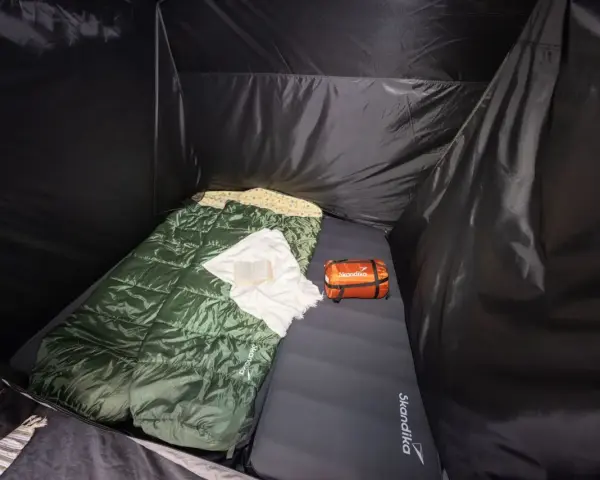 Schlafsack und Kissen auf der Isomatte, welche Seite oben ist, ist klar erkennbar. Ein kleines orangefarbenes Päckchen liegt daneben.