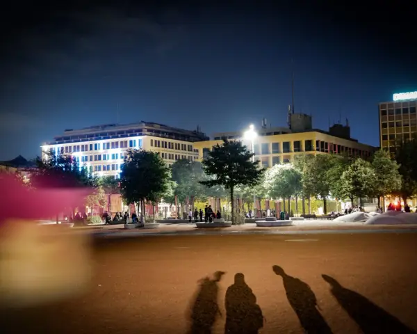 Ombre d'un mineur dehors la nuit, projetées sur une place éclairée par des bâtiments modernes.