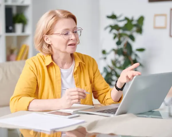 Kobieta w okularach i żółtej koszuli pracuje przy laptopie, trzymając długopis. To jej dodatek funkcyjny do pracy zdalnej.