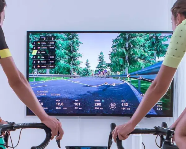 Two cyclists race virtually on a scenic road using indoor cycling apps. The screen displays their stats and rankings.