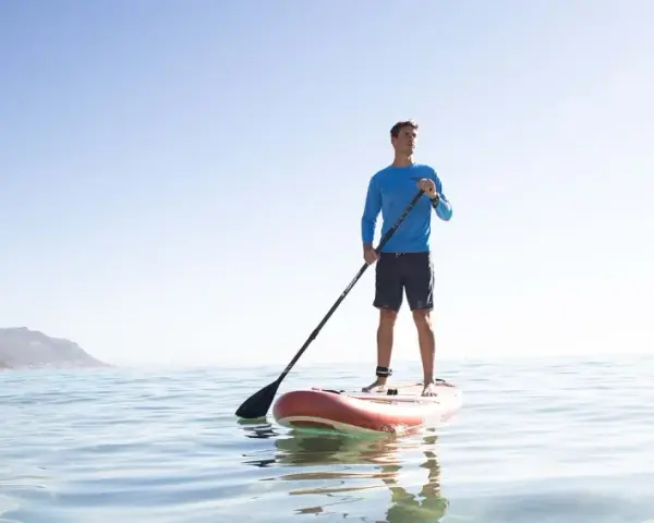 Mann steht auf SUP-Board, hält Paddel richtig. Ruhige See, bergige Küste im Hintergrund.