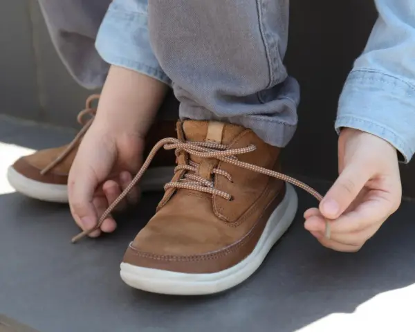 Kind lernt, seine braunen Schnürstiefel zu binden. Die Schnürsenkel sind hellbraun und gemustert.