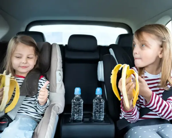 Dwie dziewczynki w fotelikach samochodowych jedzą banany. Woda i przekąski to podstawa, gdy zastanawiasz się, co zabrać na długą podróż samochodem z dzieckiem.