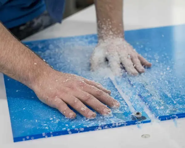 Hands guide a router, creating a clean cut on a blue plexiglass sheet, leaving a trail of white shavings. This is how you cut plexiglass with a router.