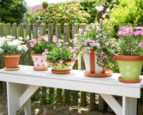 Fünf bemalte Blumentöpfe mit bunten Blumen stehen auf einer weißen Bank im Garten. Ein Blumentopf ist grün mit weißen Blumen bemalt.