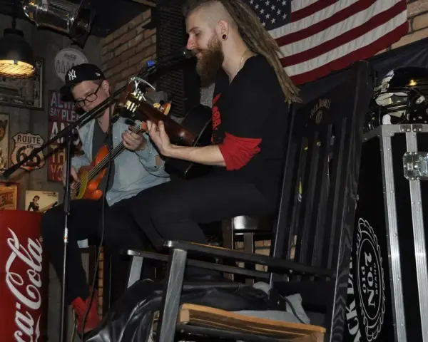 Dwóch muzyków gra na gitarach w klimatycznym wnętrzu. Jeden z nich, muzyk grający do kotleta, ma dredy i brodę.
