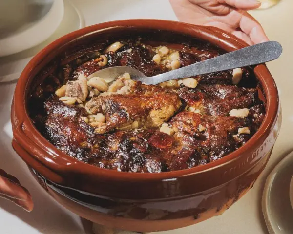 Un plat en terre cuite débordant de haricots blancs, de morceaux de viande dorée et de saucisses, prêt à être dégusté. La recette du vrai cassoulet traditionnel, un délice réconfortant.