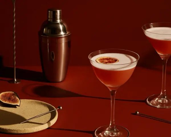 Elegancki drink z czerwonym winem, ozdobiony plasterkiem cytrusa, obok shakera i kieliszka z pianką.