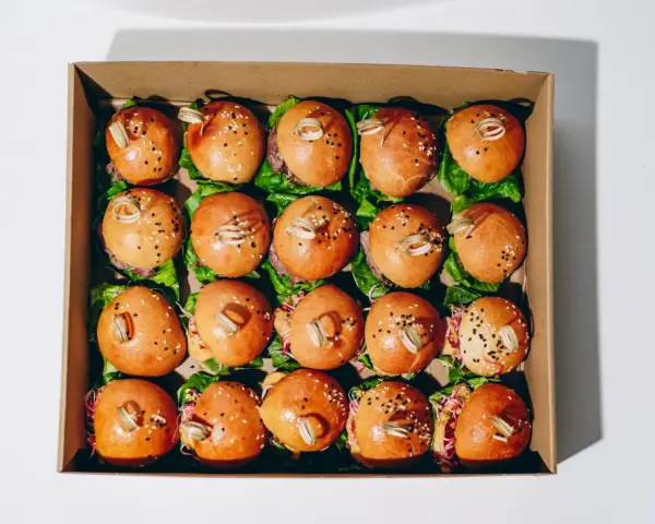 Pudełko z dwudziestoma mini burgerami na liściach sałaty, posypane sezamem i ozdobione patyczkami.