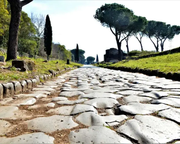 Ancient Roman cobblestone road lined with trees and ruins, leading towards the horizon.