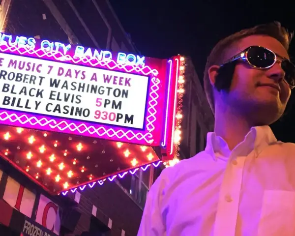 Mann im Elvis-Stil mit Sonnenbrille vor der "Blues City Band Box"-Neonreklame, die "Black Elvis" ankündigt.