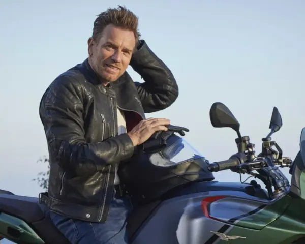 Ewan McGregor, con giacca di pelle, posa su una Moto Guzzi verde e grigia, pronto per un'avventura.