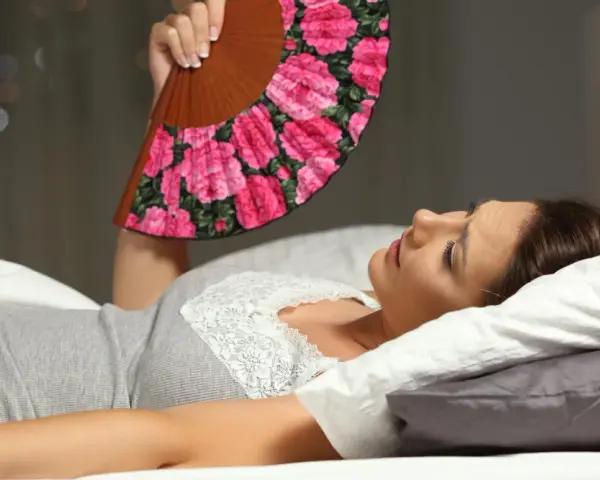Frau liegt im Bett, nass geschwitzt, fächelt sich mit einem Blumenfächer Luft zu. Die Nacht ist heiß.