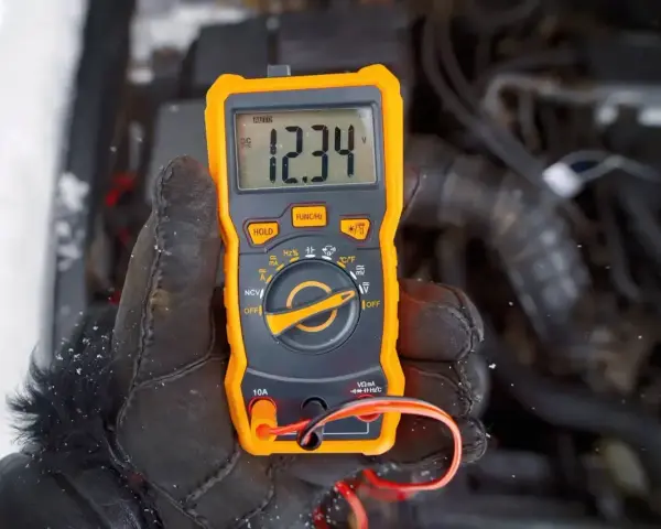 Ręka w rękawicy trzyma pomarańczowy multimetr, który pokazuje 12.34V. Auto nie odpala, akumulator sprawny, problem leży gdzie indziej.
