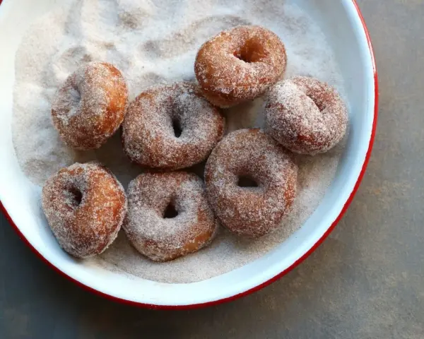 Små, nybakade donuts som rullats i socker och kanel ligger i en vit skål med röd kant.