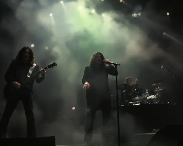 Un groupe de metal sur scène, avec un guitariste à gauche, un chanteur au centre, et un batteur à droite. La scène est enfumée, éclairée par des projecteurs.