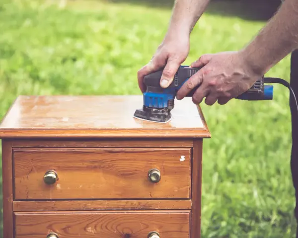 Un homme utilise une ponceuse pour le décapage mécanique d'une commode en bois, préparant le meuble pour une nouvelle finition.