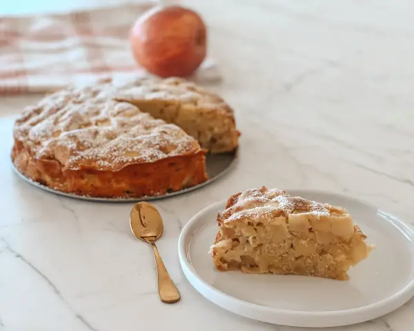 Une part de **crépiau aux pommes** saupoudrée de sucre glace, à côté d'un gâteau entier et d'une pomme. Une cuillère dorée est posée sur l'assiette.