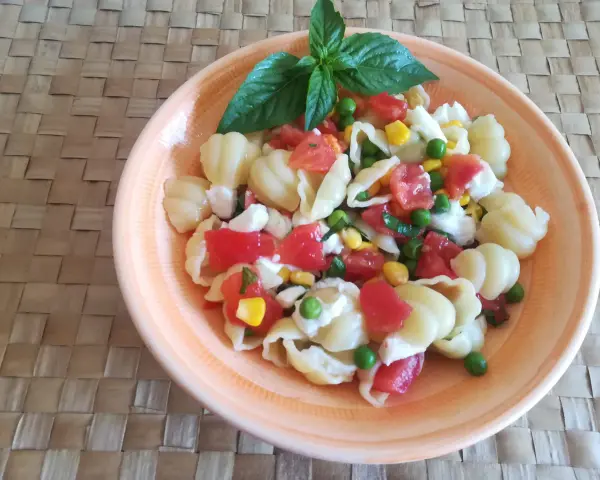 Un'appetitosa insalata di pasta vegetariana con pomodorini, piselli, mais e mozzarella, guarnita con basilico fresco.