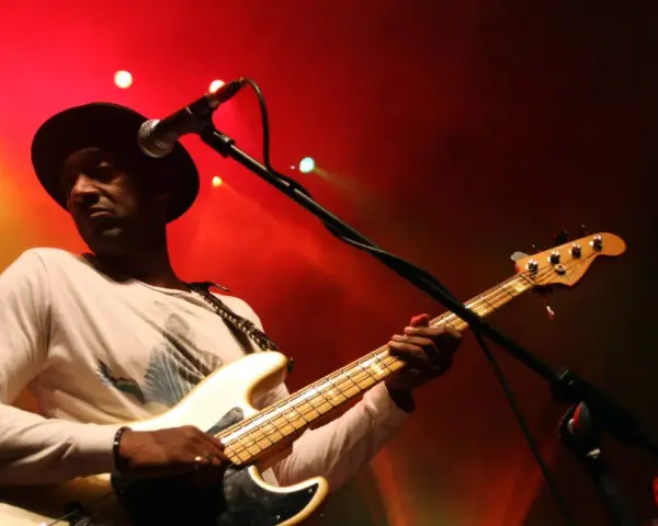 Marcus Miller w czarnym kapeluszu gra na białej gitarze basowej na tle czerwonych świateł scenicznych.