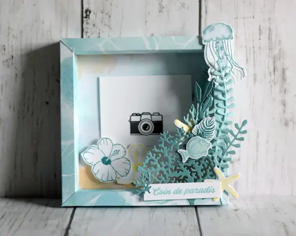 Un cadre photo en bois décoré sur le thème marin avec une méduse, des poissons, des fleurs et une petite caméra.