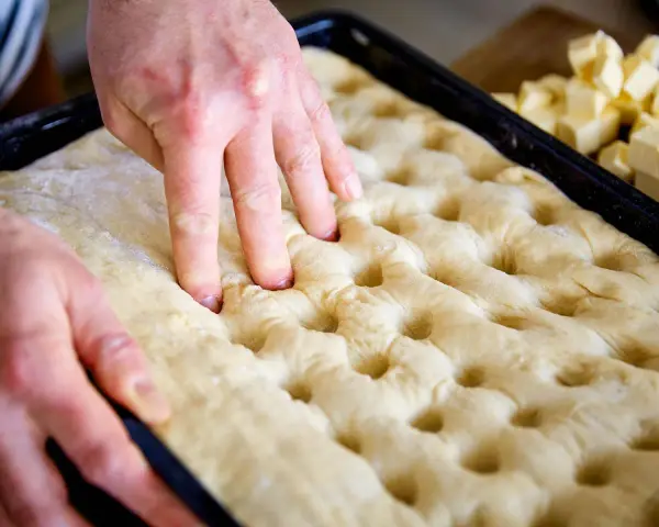 Hände drücken Vertiefungen in den Teig für ein Butterkuchen Rezept. Würfel aus Butter liegen bereit.