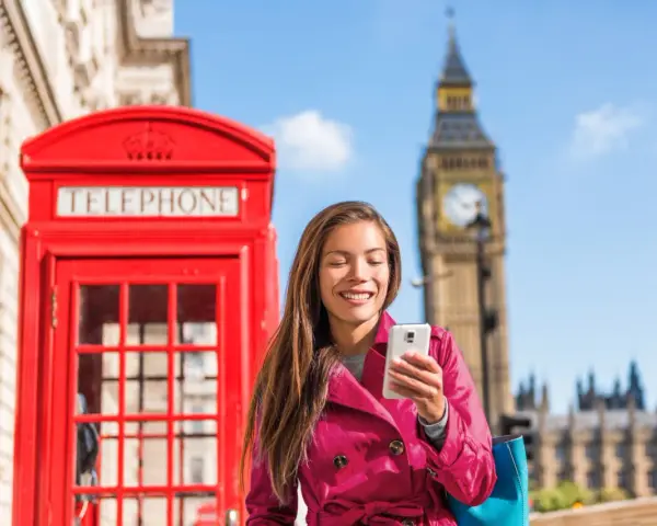 Uśmiechnięta kobieta z telefonem przed czerwoną budką telefoniczną i Big Benem. Pyta, czy w UK jest roaming, bo chce zadzwonić do domu.