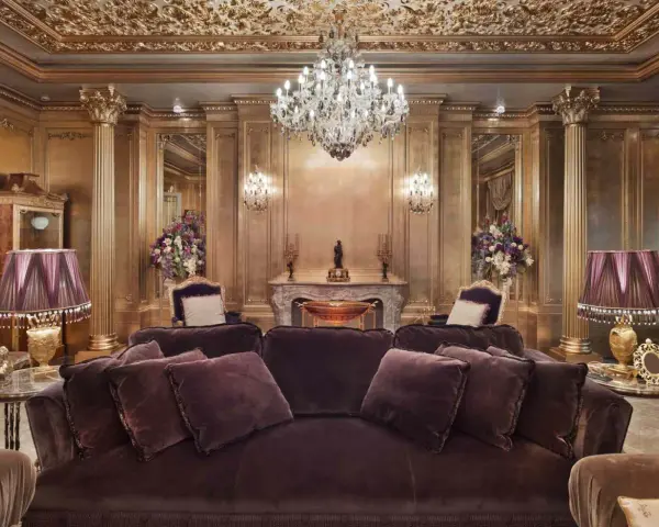 Salón lujoso con sofás de terciopelo morado, lámparas doradas y un candelabro de cristal. El techo y las paredes doradas evocan el estilo barroco español.