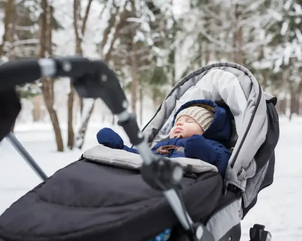 Śpiące dziecko w wózku zimą. Ciepły kombinezon, czapka i rękawiczki to podstawa, co włożyć do wózka dla noworodka zimą.