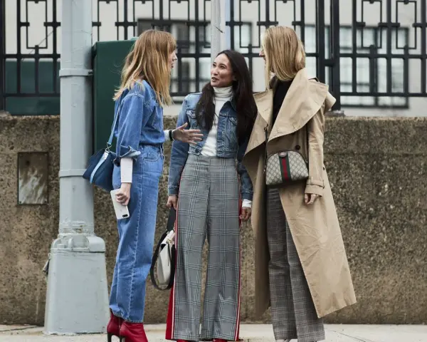 Tres mujeres conversan, una con un traje completo de **denim**, otra con chaqueta **denim** y pantalones a cuadros, y la tercera con un abrigo beige y pantalones a cuadros.