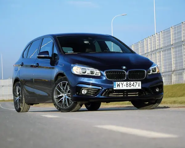 Ciemnoniebieski BMW van na tle jasnego nieba i barier dźwiękochłonnych.