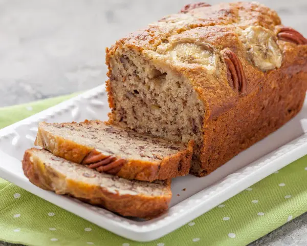 Pyszny chlebek bananowy z 2 bananów, z orzechami pekan i plasterkami banana na wierzchu, na białej paterze.