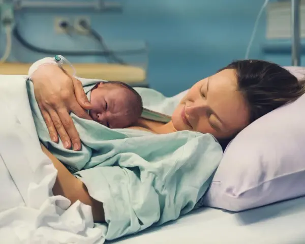 Eine Mutter hält ihr Neugeborenes im Arm, glücklich und erschöpft. Die Szene zeigt, wie lange nach Geburt im Krankenhaus die erste Bindung entsteht.