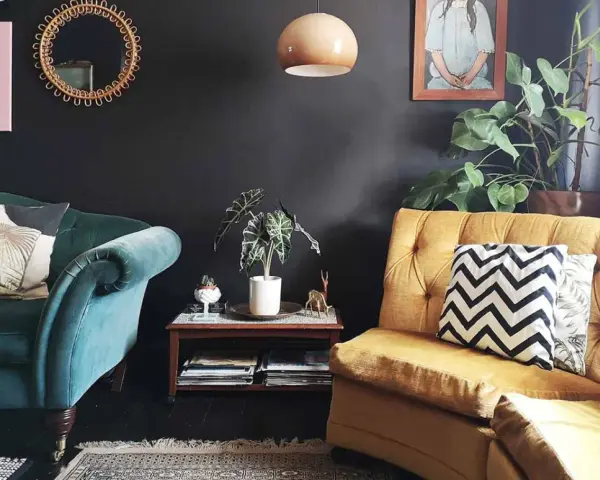 Habitación con poca luz, sofás de terciopelo verde y mostaza, cojines decorativos, plantas y arte en la pared.