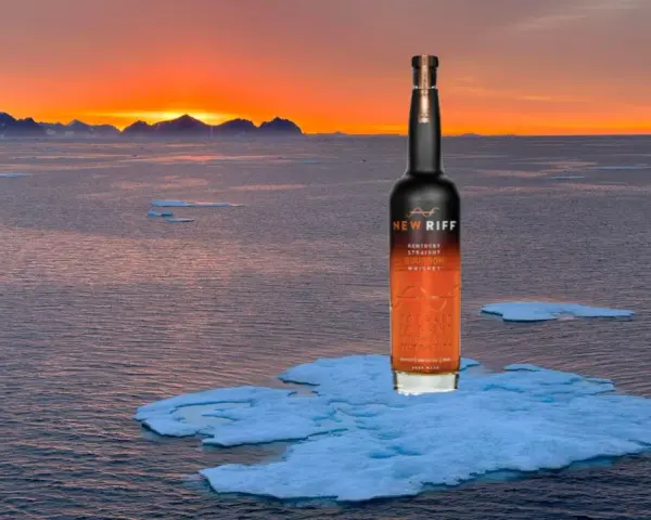 Eine Flasche New Riff Bourbon Whiskey steht auf einer Eisscholle im Meer. Der Sonnenuntergang taucht die Berge am Horizont in warmes Licht. Ein Moment der Ruhe für den besten Whisky der Welt.