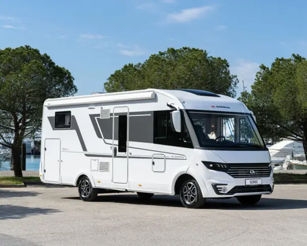 En vit Adria Sonic husbil, den lyxigaste husbilen, parkerad vid en marina med båtar och träd i bakgrunden.
