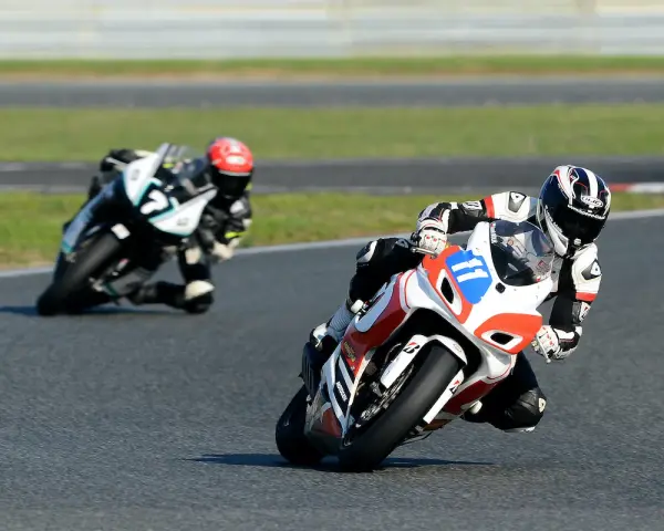 Dwóch motocyklistów w pełnym rynsztunku ściga się na torze. Jeden z nich, z numerem 11, pochyla się nisko w zakręcie, demonstrując emocje sportu motocyklowego.
