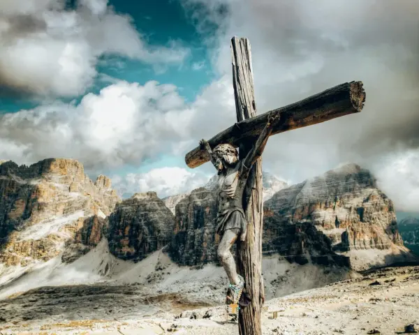 Jesus am Kreuz, eine hölzerne Figur, steht vor majestätischen Bergen unter einem dramatischen Himmel.