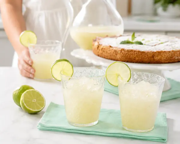 Dos vasos de granizado de limón refrescante, adornados con rodajas de lima, junto a limones frescos y un pastel.