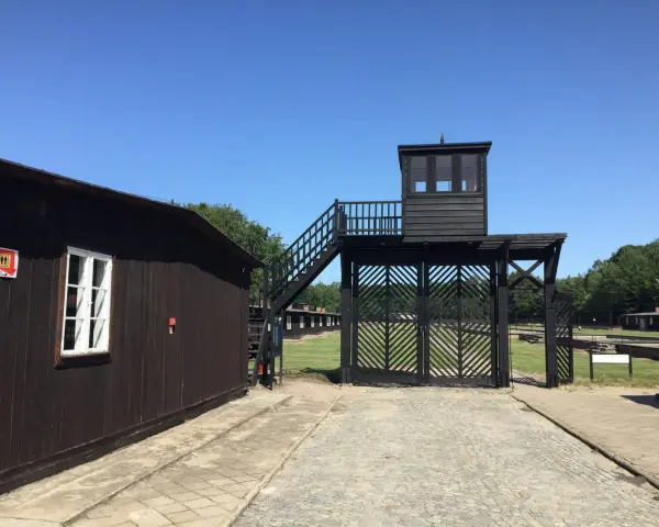 Brama i wieża strażnicza na terenie Muzeum Stutthof. Drewniane budynki i brukowany chodnik.