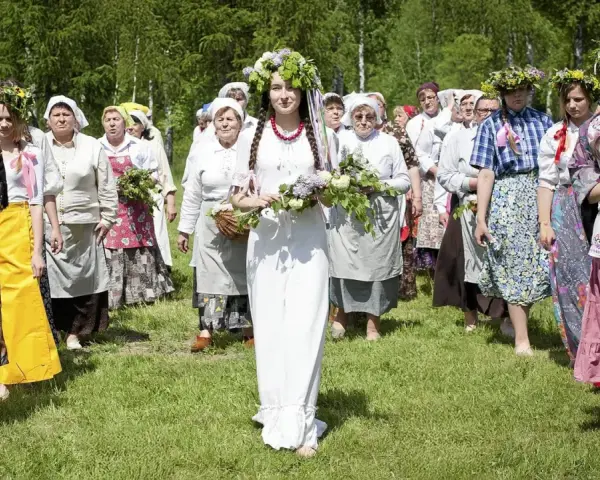 Grupa kobiet wiankach na głowach i tradycyjnych strojach idzie przez łąkę. Obraz nawiązuje do święta zielone świątki.